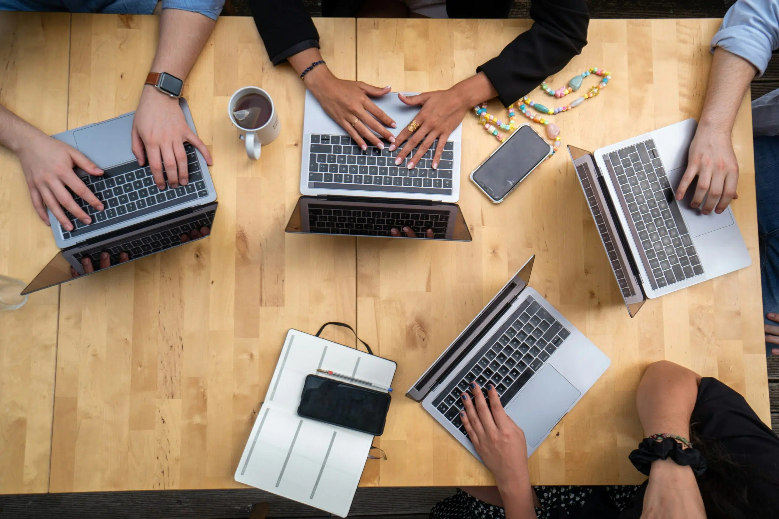 Overhead view of multiple hands working on laptops around a wooden table with coffee and notebooks.